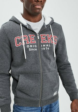 Grijze zip-up hoodie met witte trekkoorden, voorzakken en een pluizige voering. Bevat rode en witte tekst: "CREEKS Original Since 1974."