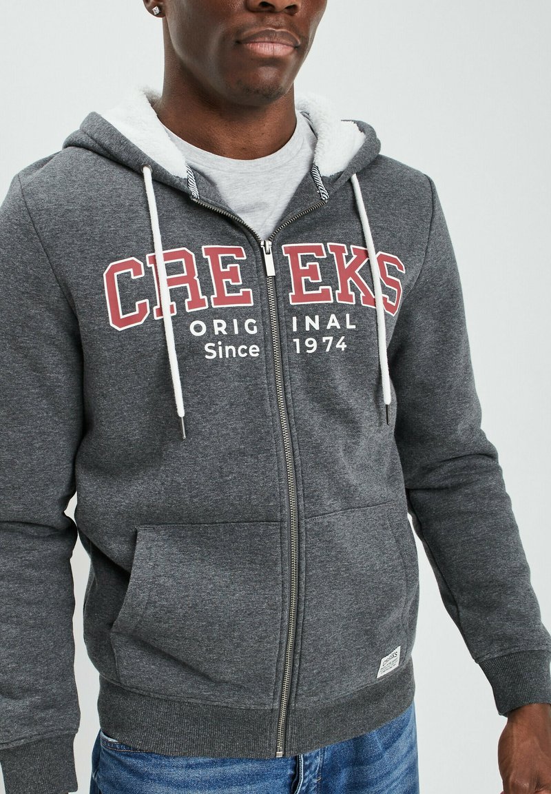 Sudadera gris con cremallera, cordones blancos, bolsillos frontales y forro de felpa. Presenta texto en rojo y blanco: "CREEKS Original Desde 1974."