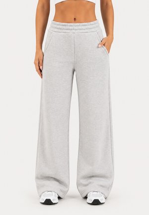 Femme portant un pantalon de jogging ample taille haute gris clair avec une ceinture élastique et des baskets blanches, debout avec une main dans la poche.