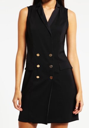Femme portant une robe noire sans manches à double boutonnage avec des boutons dorés et des poches à rabat, visible des épaules jusqu'au milieu de la cuisse.