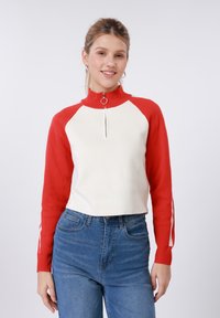 Λευκό και κόκκινο πλεκτό πουλόβερ με φερμουάρ και ψηλό γιακά, με κόκκινα μανίκια και λευκές πλαϊνές ρίγες, σε συνδυασμό με μπλε τζιν τζιν.