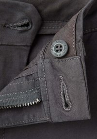 Gros plan sur un tissu gris avec un bouton marqué MNG près d'une boutonnière cousue et d'une fermeture éclair partiellement ouverte.