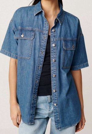 Persoon draagt een blauw denim overhemd met korte mouwen over een zwart topje en lichtblauwe jeans, staand met ontspannen armen langs het lichaam.
