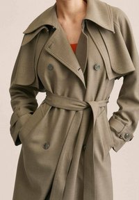 Abrigo trench color beige con diseño de doble botonadura, cuello ancho y cintura con cinturón. Hecho de tela suave con detalles de botones.