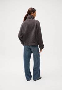 Kvinde med langt hår i en hestehale, iført en ternet jakke med høj krave og løse blå jeans, vendt væk mod en ensfarvet baggrund.