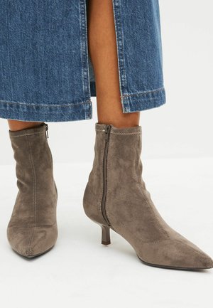 Vrouw die taupekleurige suède laarzen tot halverwege de kuit draagt met zijritsen en lage hakken, gecombineerd met blauwe denim wijde pijpen broek.