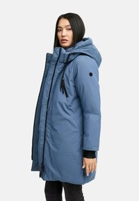 Donna che indossa un lungo cappotto invernale blu imbottito con cappuccio, in piedi di profilo su uno sfondo chiaro e semplice.