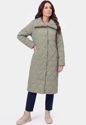 Frau mit langen braunen Haaren, die einen langen, gestepp-ten hellgrünen Mantel, marineblaue Hose und beige Schnürstiefel trägt und mit einer Hand in der Hüfte steht.