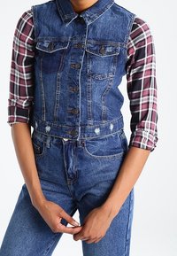 Jeansvest met kraag, voorkantzakken en knoopsluiting, over een langemouw, geruit shirt in donkerrood, wit en zwart.