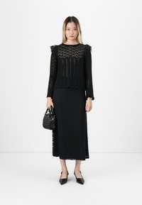 Kvinne iført en svart blonde bluse med lange ermer og matchende lang skjørt, som holder en liten svart håndveske og har på seg svarte spisse sko.