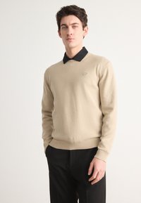 Beige kereknyakú pulóver, puha anyagból készült, fekete galléros inggel alatta, és egy kör alakú logóval a bal mellen.