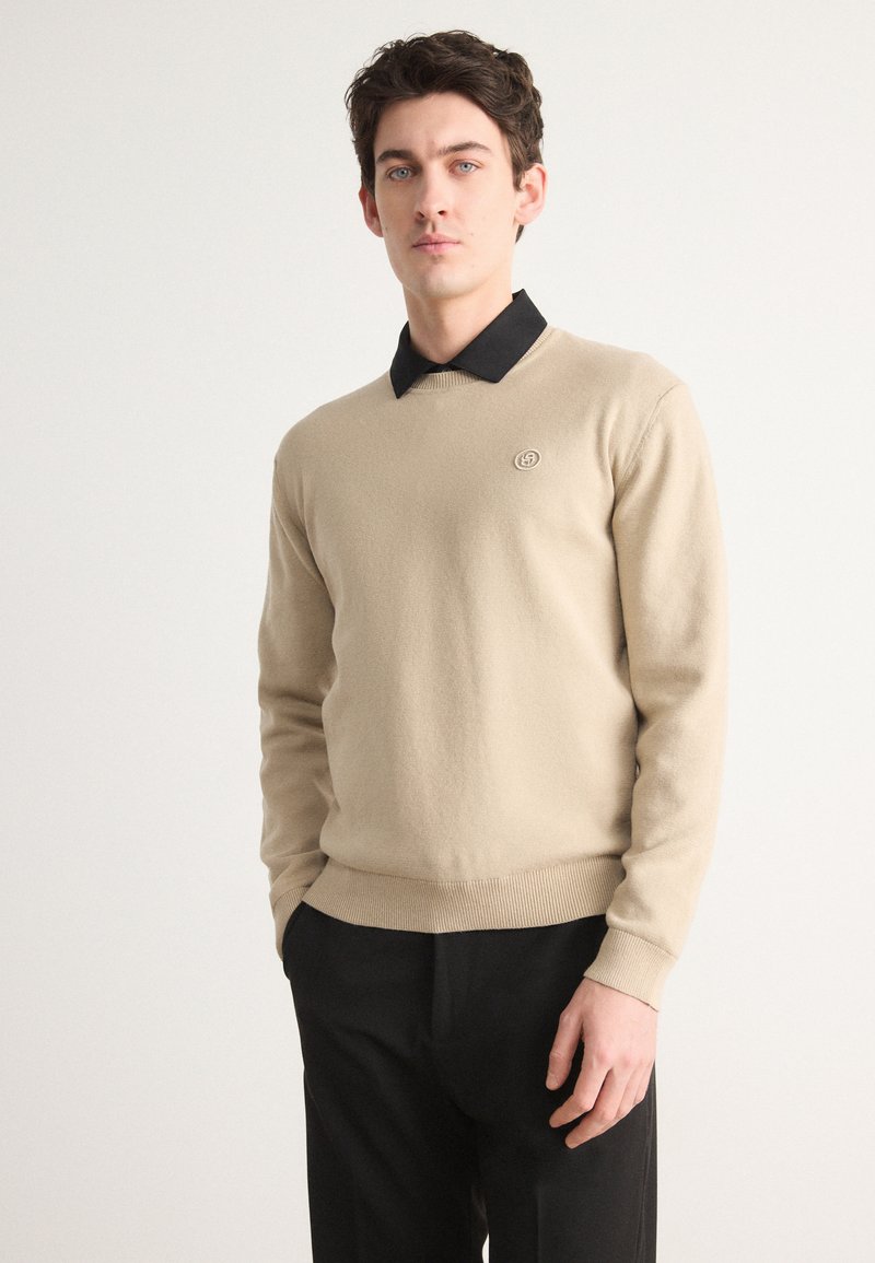 Beige kereknyakú pulóver, puha anyagból készült, fekete galléros inggel alatta, és egy kör alakú logóval a bal mellen.