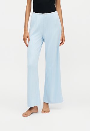 Femme portant un pantalon lounge large côtelé bleu clair avec taille élastique et petit nœud, debout pieds nus sur fond blanc.