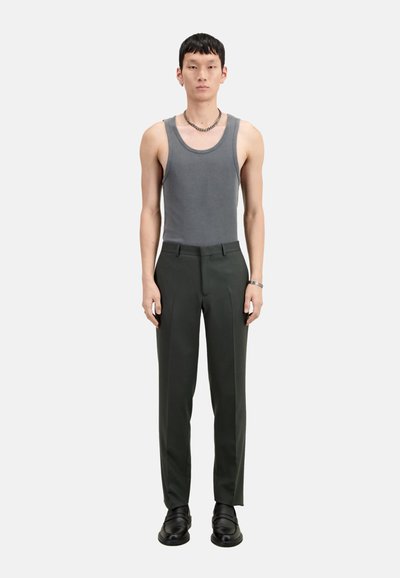 Graues Tanktop, taillierte dunkelgrüne Hose, schwarze klobige Schuhe und ein silbernes Armband. Das Modell steht aufrecht vor einem weißen Hintergrund.