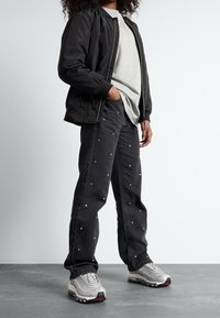 Černá bomber bunda přes šedé tričko, doplněná černými džínami se stříbrnými cvoky a šedými teniskami. Pozadí je světle šedé.