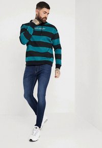 Teal och svart randig sweatshirt med huva, kombinerad med blå slim-fit jeans och vita sneakers. Har en logotyp på bröstet.