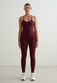 Burgundowy sportowy top ze szwami i cienkimi ramiączkami, w komplecie z pasującymi legginsami wysoko w talii; całość dopełniają białe buty sportowe.