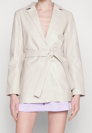 Blazer in pelle beige con cintura e ampi revers indossato sopra pantaloncini lilla chiaro arrotolati su una persona in piedi.