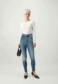 Modre jeans hlače z visokim pasom, z dvema strganima predeloma, v kombinaciji z belo dolgo majico in črnimi čevlji s koničastim nosom. Drži črno torbico.