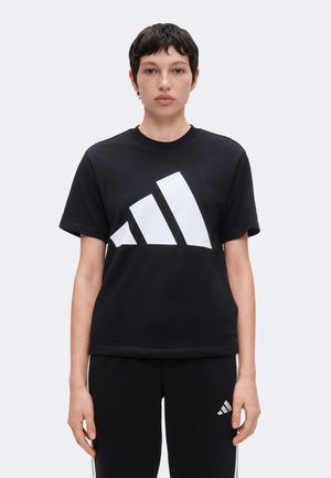 Jeune personne aux cheveux courts et foncés, portant un T-shirt noir et un pantalon avec de larges bandes blanches en diagonale sur le T-shirt et un petit logo sur le pantalon.