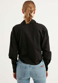 Schwarz langärmliges Hemd mit Kragen, lockerer Passform und Knopfmanschetten. Kombiniert mit hellblauen, eng anliegenden Jeans mit Gesäßtaschen.