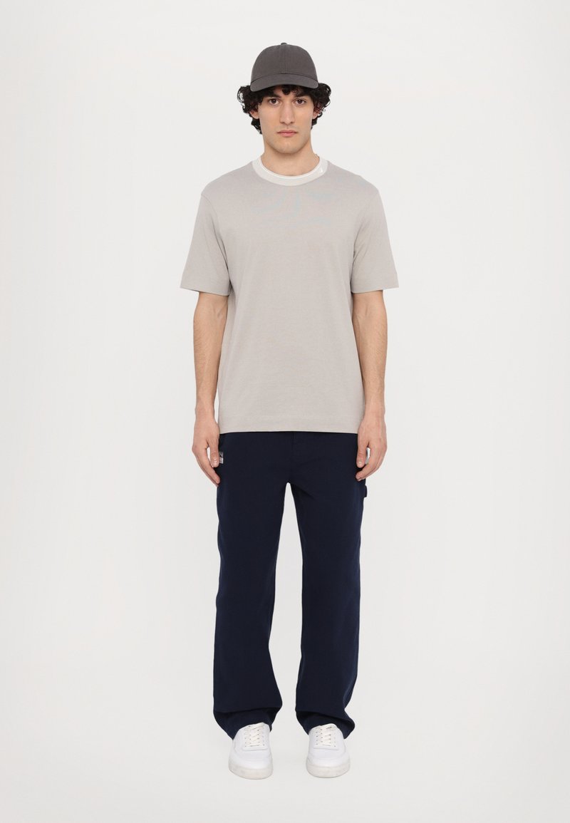 Camiseta de manga corta gris claro con cuello redondo blanco, combinada con pantalones azul marino de corte holgado y zapatillas blancas, complementada con una gorra gris.