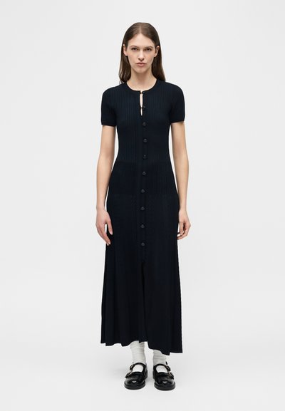 Γυναίκα που φοράει μαύρο, μίντι φόρεμα με κοντά μανίκια και κουμπιά, λευκές κάλτσες και μαύρα παπούτσια Mary Jane, στέκεται μπροστά σε απλό λευκό φόντο.