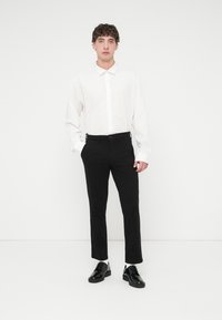Chemise blanche à boutons, pantalon noir sur mesure, et chaussures noires brillantes. La chemise a des manches longues ; le pantalon est ajusté et à coupe droite.