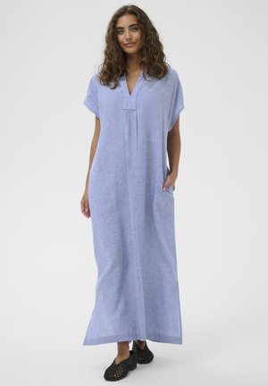 Göndör hajú nő laza, világoskék, bokáig érő ruhát visel, rövid ujjúval és fekete szövött belebújós cipővel.