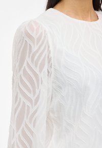 White blouse with textured wavy pattern, featuring semi-transparent long sleeves and a round neckline. Soft, lightweight fabric.