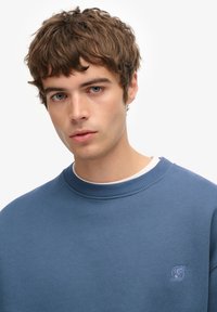 Blauwe sweatshirt met een geribde halslijn en subtiele logo-detail. Gemaakt van een zacht materiaal, heeft het een ontspannen pasvorm en een gladde textuur.