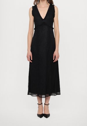 Femme portant une robe midi noire sans manches avec un décolleté en V profond et une bordure à volants, associée à des escarpins noirs à bout pointu et à brides autour de la cheville.