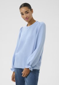 Blouse à manches longues bleu clair en tissu texturé, avec un col rond et des poignets de manches froncés. Associée à un jean en denim foncé.
