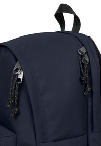 Námně modrý batoh s hladkým povrchem, se stříbrnými detaily a černými akcentními šňůrkami na zavírání. Obsahuje horní držadlo pro nošení.