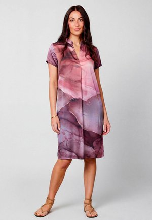 Femme debout, portant une robe courte à manches courtes avec un motif abstrait rose et violet, associée à des sandales à brides beige.