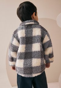 Enfant de dos portant une veste en peluche gris et blanc à carreaux et un pantalon foncé dans un environnement intérieur minimal.