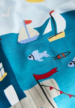 Tekstilė su ryškiai mėlynu fonu, puošta išsiuvinėtais jachtų, žuvų ir raudonos vėliavos motyvais, pabrėžianti jūrinę temą.