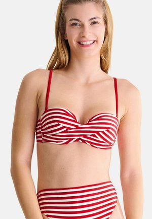 Jeune femme souriante, portant un bikini rayé rouge et blanc avec un haut bandeau torsadé et un bas taille haute, posant devant un fond uni.