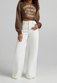 Sweatshirt corto marrone con testo bianco, abbinato a jeans bianchi a vita alta e gamba larga. Entrambi realizzati in cotone con una texture liscia.