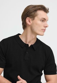 Polo noir en tissu texturé avec col, deux boutons au niveau du col et manches courtes, présenté sur un mannequin de profil.