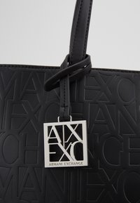 Svart totebag med preget logo-design, glatt tekstur, og et sølvfirkanthengende vedhenget med merkenavnet "Armani Exchange."