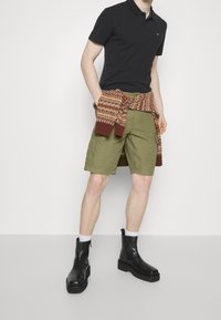 Polo nero, pantaloni cargo verde oliva, un maglione a motivo marrone legato in vita e stivaletti neri con suola piatta spessa.