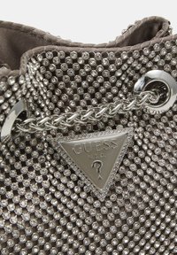 Borsa grigia con dettagli metallici, decorata con numerosi strass rotondi e una placca triangolare in metallo con logo. Dettaglio in catena argentata visibile.