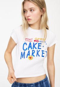 Hvid kortærmet t-shirt med farverig grafisk tekst, der står "KAGEMARKED" samt yderligere tekst. Croppet snit med rund halsudskæring.