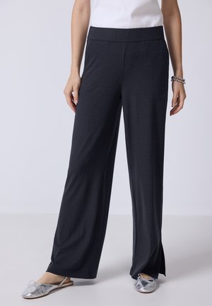 Black ribbed wide-leg pants with a high elastic waistband, featuring side slits at the hem, paired with metallic silver slip-on shoes.