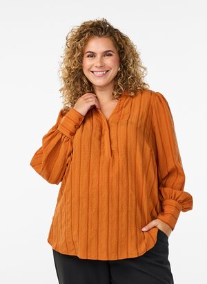 Smilende kvinde med krøllet hår iført en orange langærmet bluse og sorte bukser, stående med den ene hånd i lommen.