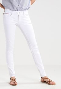 Jeans skinny blancos con una cintura de media altura, cierre con botón y diseño clásico de cinco bolsillos. Textura suave con un acento rojo en el bolsillo.