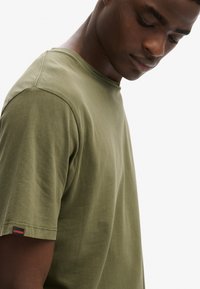 Oliwkowozielona bawełniana koszulka z okrągłym dekoltem, krótkimi rękawami i subtelną metką z logo na dole. Miękka w dotyku i o luźnym kroju.
