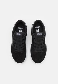 Μαύρα σουέτ αθλητικά παπούτ�σια με λευκό λογότυπο στο πλάι, με επίπεδες και κορδόνια και μαλακούς γιακάδες. Η σόλα είναι λευκή και με υφή για πρόσφυση.