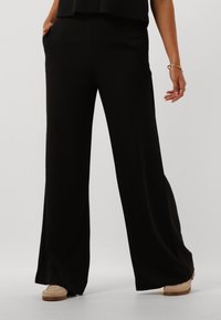 Pantaloni neri a gamba larga realizzati in un tessuto liscio, con vita alta e tasche laterali. Abbinati a calzature di colore chiaro.